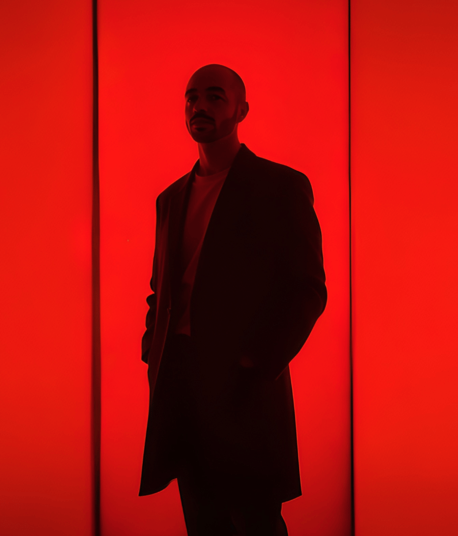 Das Bild zeigt eine Silhouette einer Person vor einem roten Hintergrund. Die Person trägt einen langen Mantel. Die starke Beleuchtung erzeugt einen dramatischen Kontrast und hebt die dunkle Silhouette gegen den kräftigen roten Hintergrund hervor.