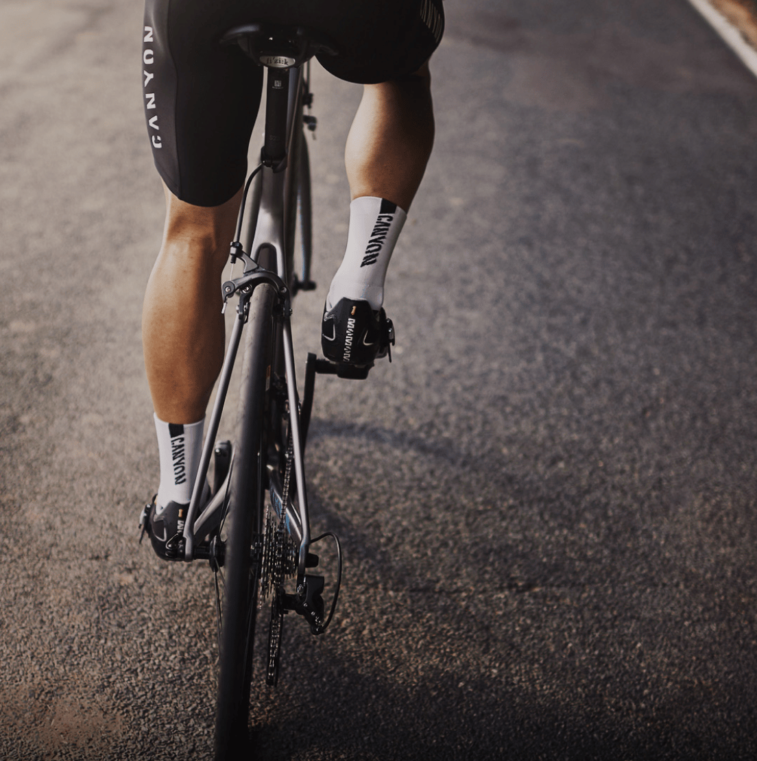 Das Bild zeigt die Beine eines Radfahrers, der auf einem Rennrad fährt. Die Beine sind in Radlerhosen gekleidet, und das Rennrad hat einen schwarzen Rahmen. Der Fahrer fährt auf einer Straße, deren Oberfläche leicht uneben ist.