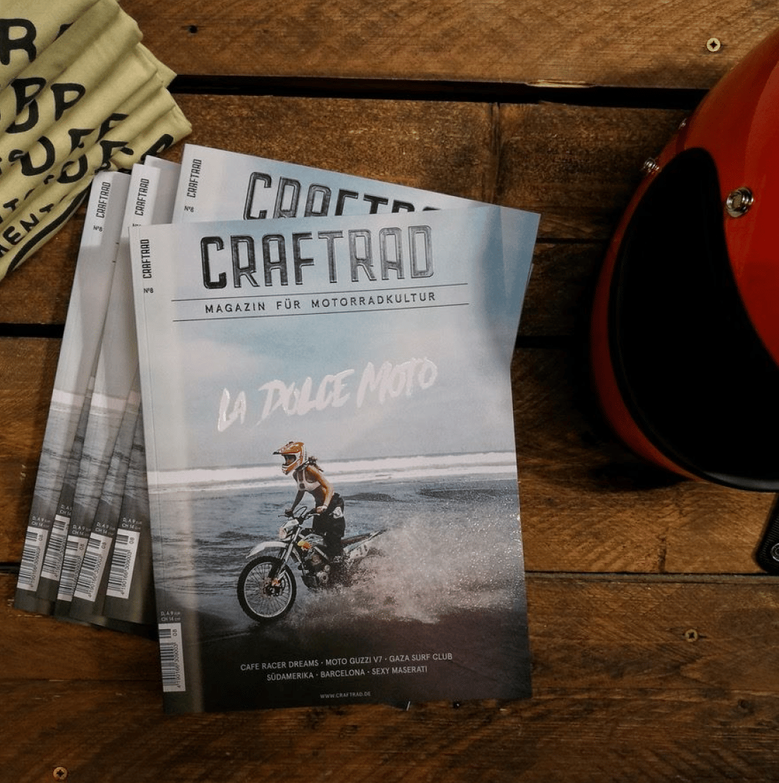 Das Bild zeigt mehrere Exemplare des Magazins “Craftrad – Magazin für Motorradkultur” auf einem Holztisch. Die Titelseite zeigt eine Person auf einem Motorrad, die am Strand fährt, mit dem Schriftzug “La Dolce Moto”. Rechts im Bild liegt ein roter Motorradhelm, links sind Stoffartikel zu sehen.