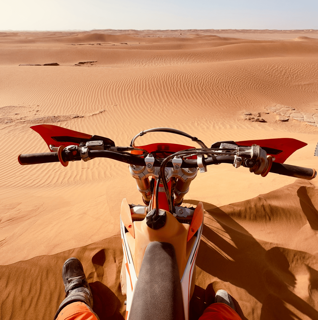 Das Bild zeigt einen Blick von oben auf ein Motorrad, das in einer weiten Wüstenlandschaft fährt. Der Vorderreifen des Motorrads ist in den Vordergrund gerückt, und der Fahrer ist nicht zu sehen. Die Wüste ist in einem warmen Braunton gefärbt und zeigt eine sanfte, gewellte Textur. Das Bild vermittelt einen Eindruck von Freiheit und Abenteuer.