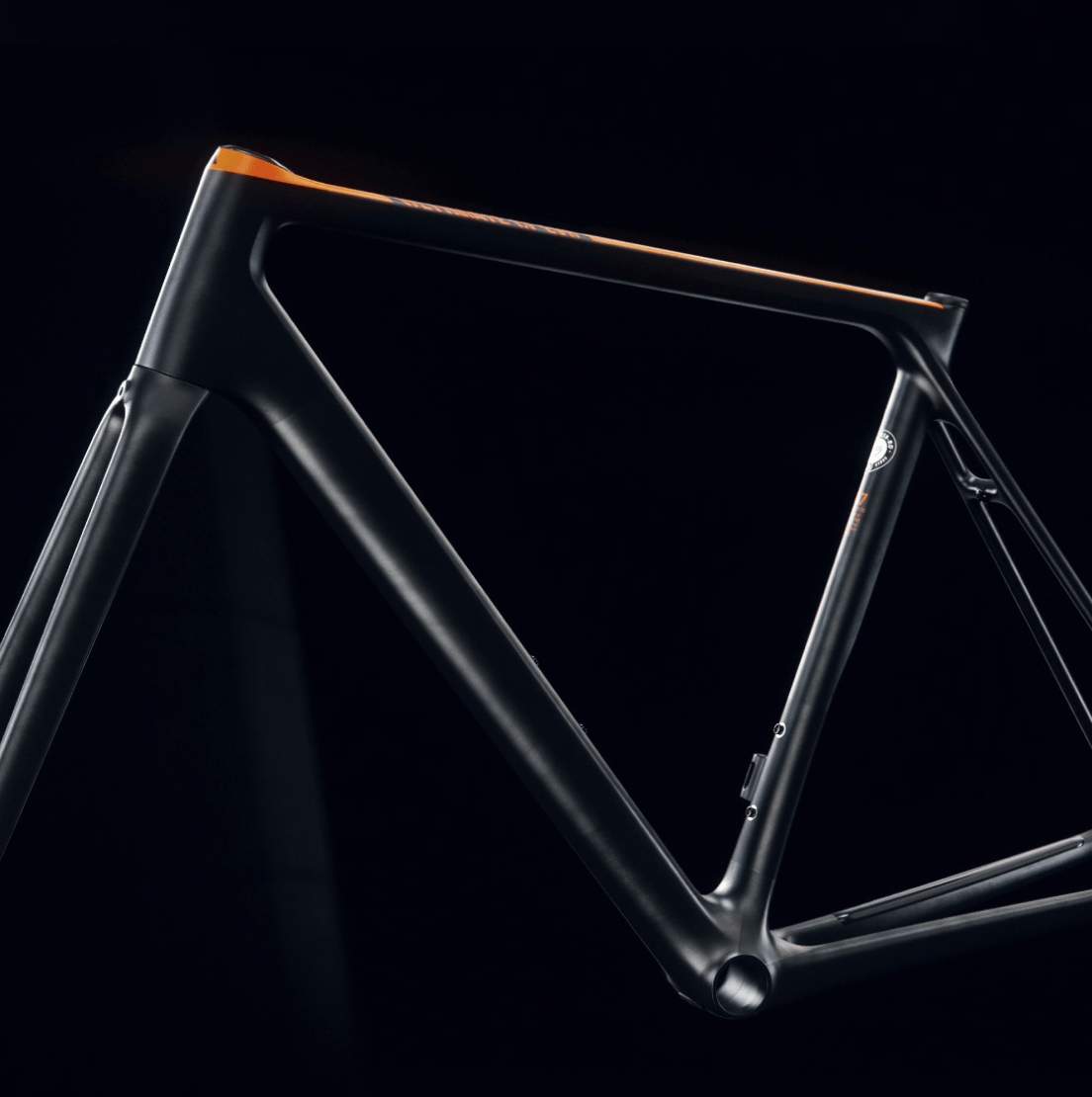 Das Bild zeigt den Rahmen eines modernen, aerodynamischen Fahrrads in mattschwarzem Design mit orangefarbenen Akzenten. Der Rahmen steht vor einem dunklen Hintergrund, der die klaren Linien und das hochwertige Material betont.